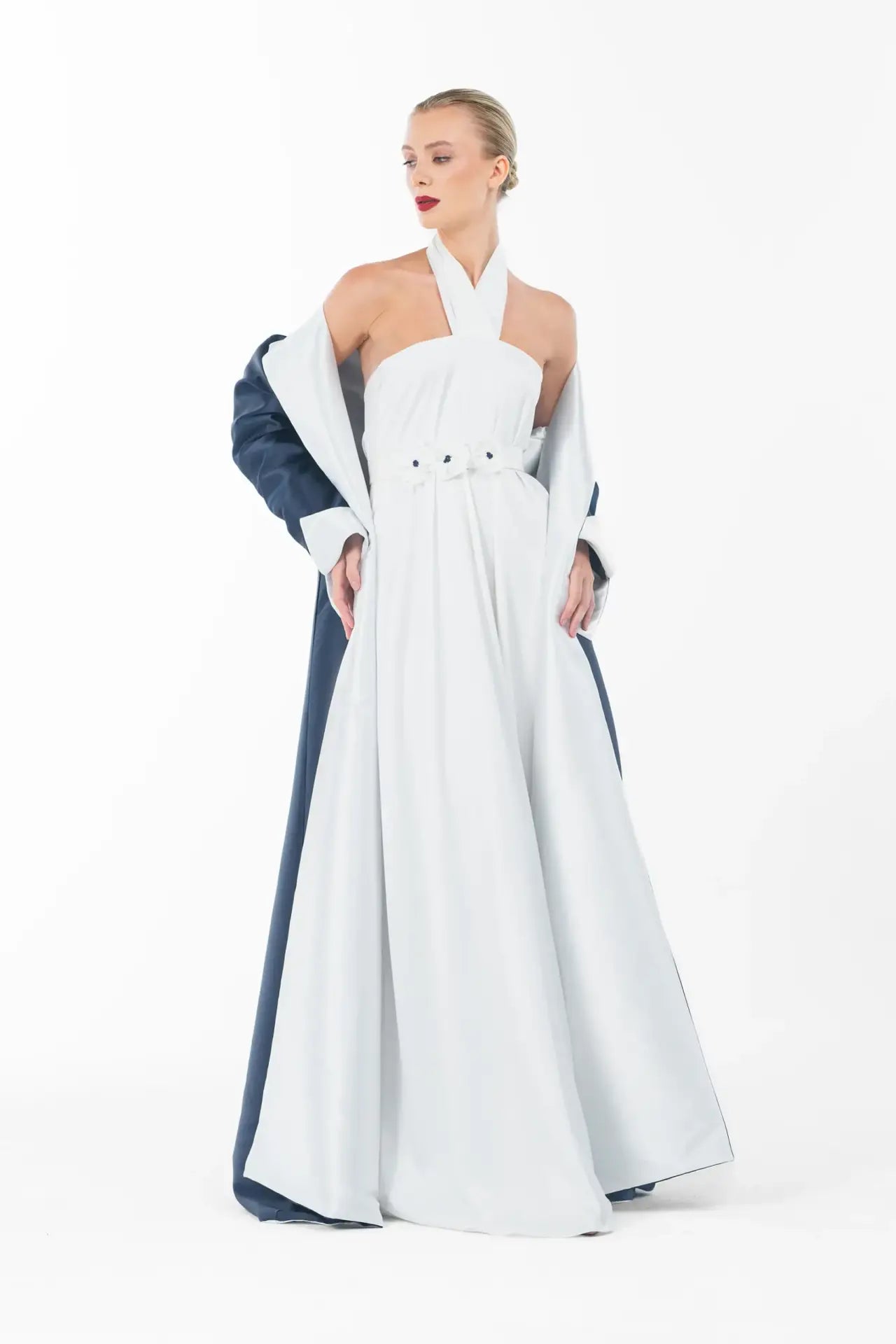 Side pose of Ava Set Artista Ramadan 2025 highlighting the halter neckline and belted waist of the white gown under the navy coat.