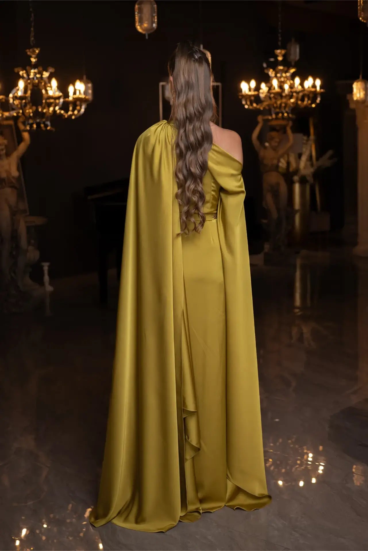 Back view of Verite Majestique PF2025 Ambre Dress in olive green satin, showcasing long draped cape and elegant one-shoulder silhouette.