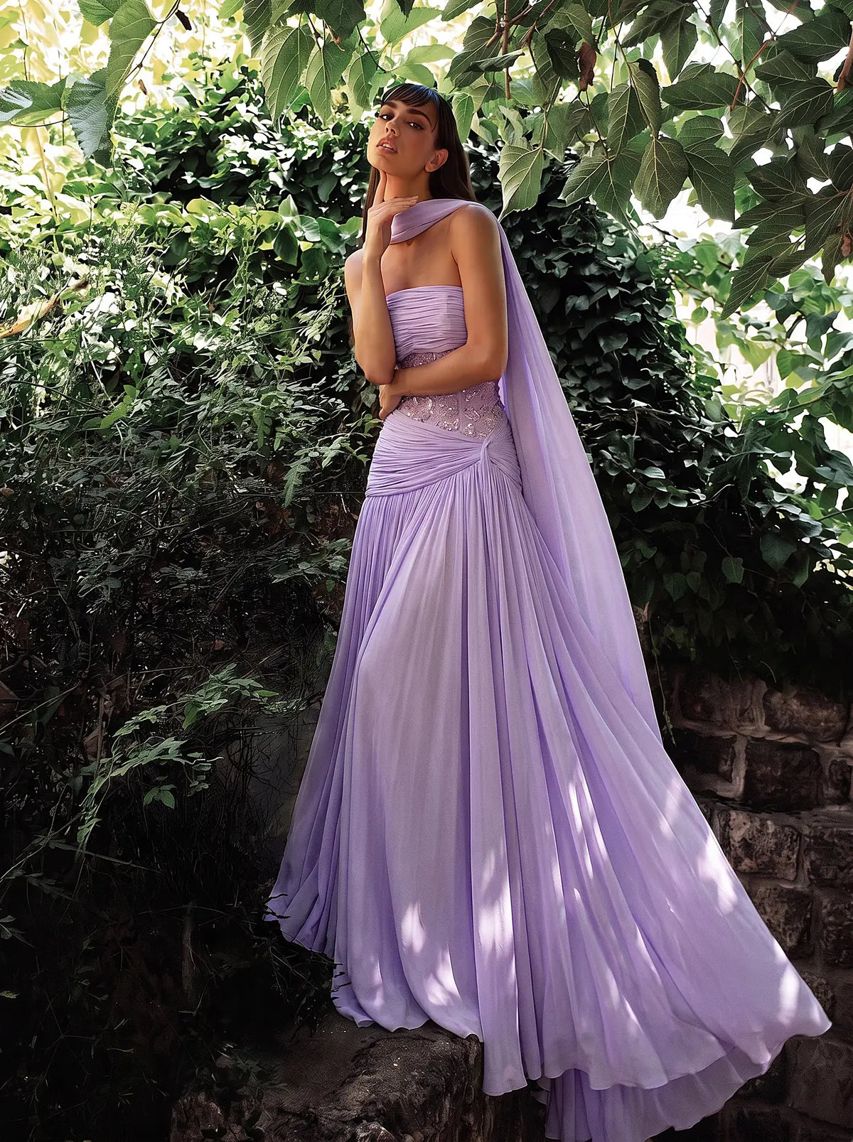 MOOEi model wearing a flowing lilac gown against green foliage.