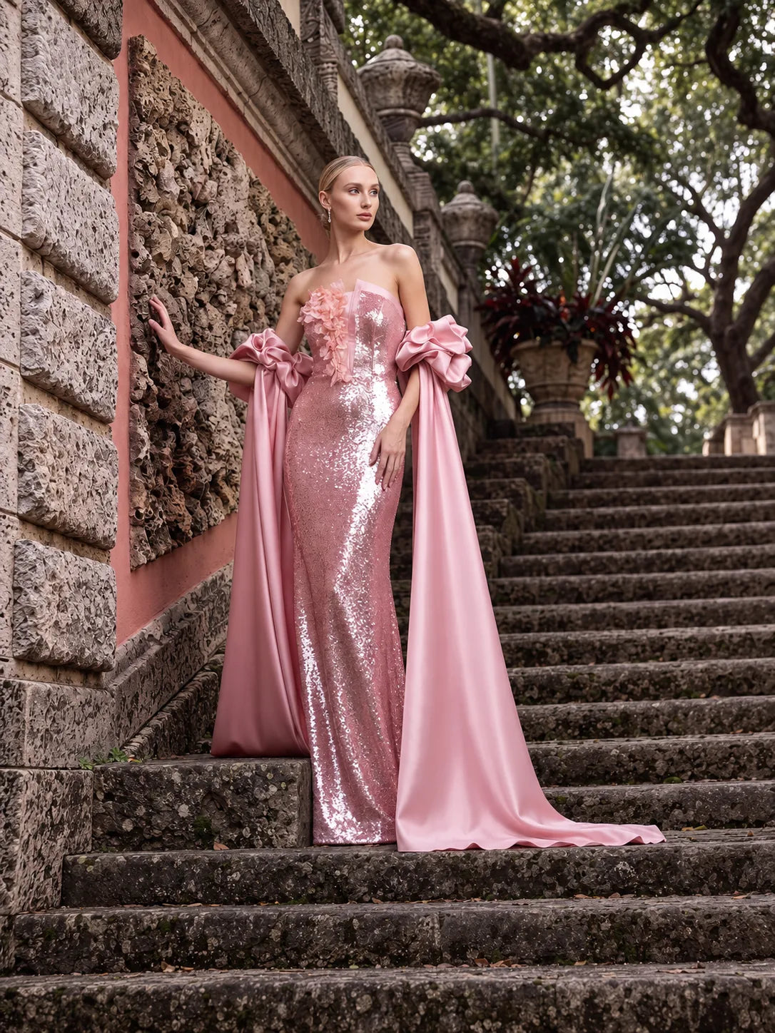 Helen of Troy by Maison MOOEi: pink sequin evening gown with sculptural satin bows and flowing cape panels on a stone stairway.